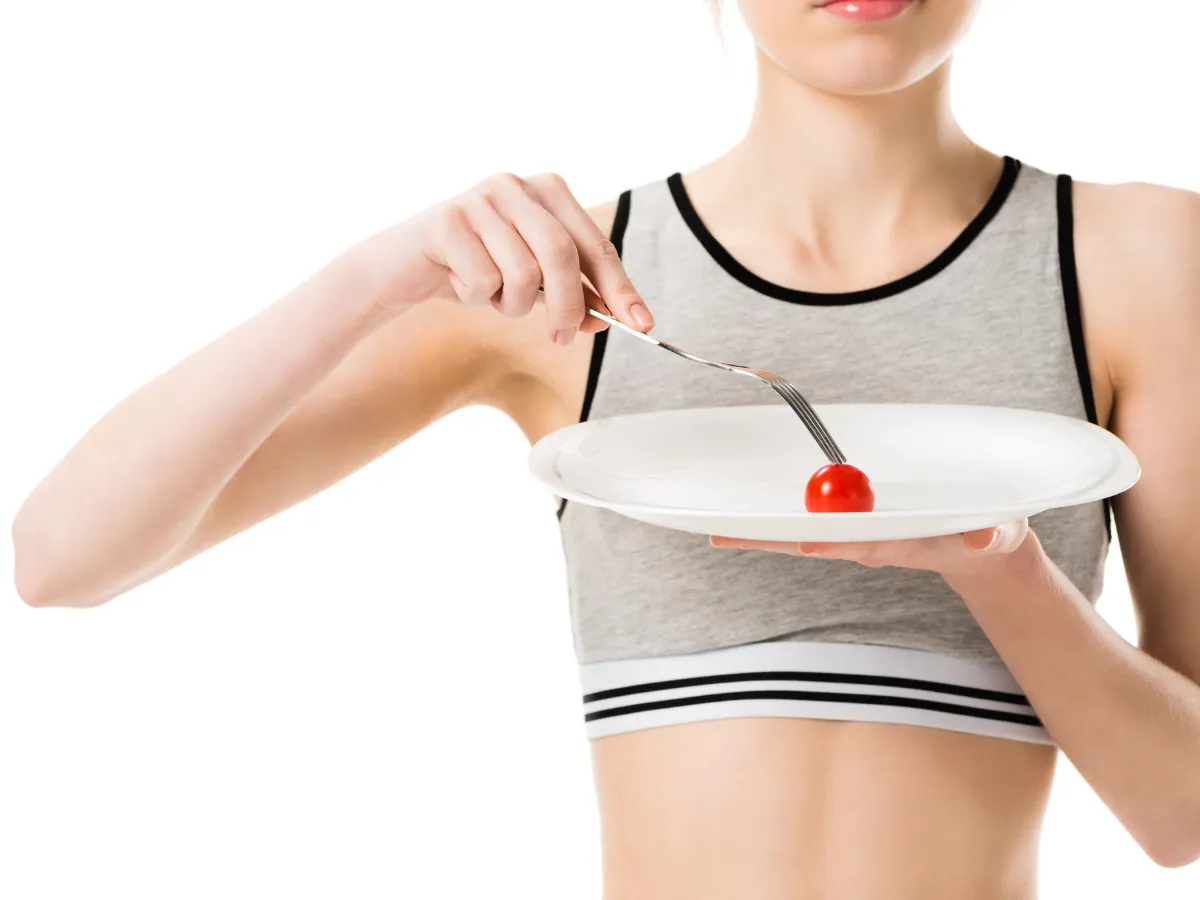 A fit young woman in a sports bra holding a white plate with a single cherry tomato, emphasizing extreme portion control or restrictive dieting.